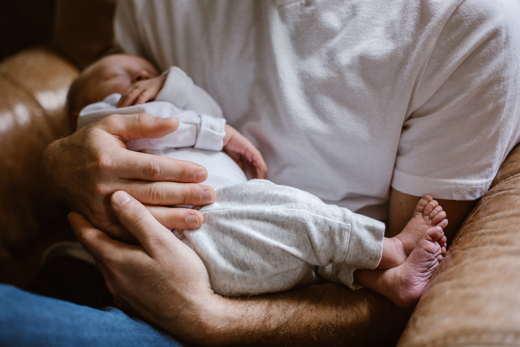 New father's hands and arms cradling his baby boy in a leather chair by the window.